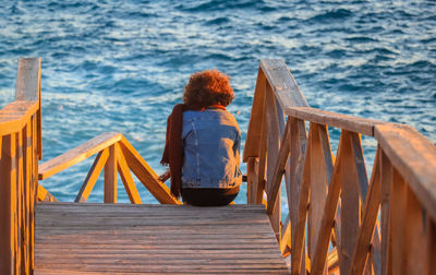 Rear view of boy sitting on wooden railing against sea