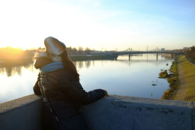 Rear view of woman looking at river while standing against sky during sunset