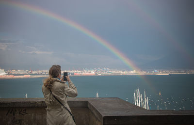 Rear view of woman photographing double rainbow and lake