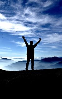 Silhouette man with arms raised standing on mountain against sky
