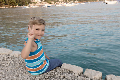 Cute boy showing victory sign while sitting on a pier at the beach.