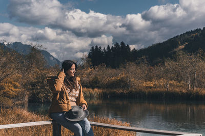 Young woman sitting on fence on shore of lake in autumn