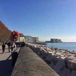 People walking on retaining wall by sea against clear blue sky