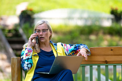 Senior woman at the garden working from home using laptop and speaking on the phone