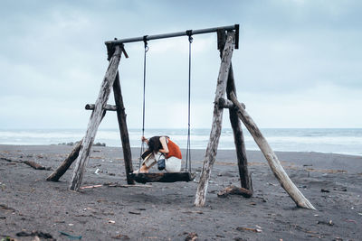 Side view of woman sitting on swing at beach against cloudy sky