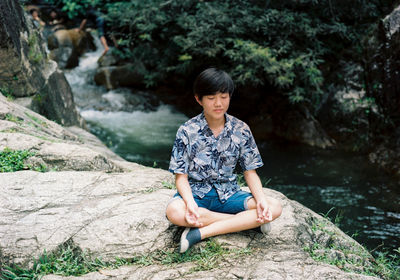 Teenage boy meditating while sitting on rock by river