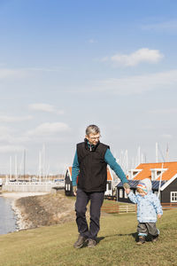 Grandfather and granddaughter walking on field against harbor in town