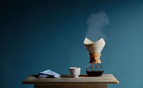 Coffee pot with cup and napkin on table against blue wall