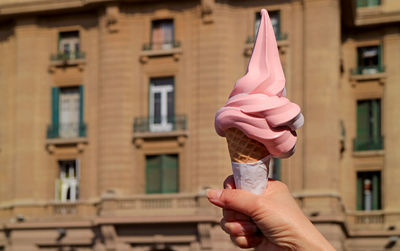 Woman's hand holding delectable pastel pink soft serve ice cream cone against vintage buildings