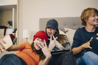 Girls taking selfie while sitting with male friend on bed at home