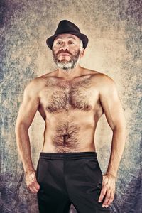 Portrait of shirtless man wearing hat standing against wall