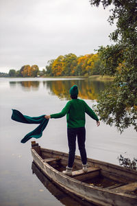 Woman in a green sweater stands with her back in an old wooden boat on a lake in autumn