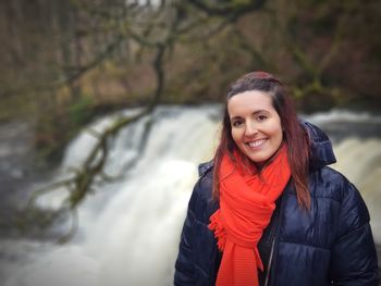 Portrait of smiling young woman wearing warm clothing while standing against waterfall