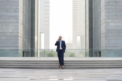 Full length of businessman talking on mobile phone while walking against office buildings