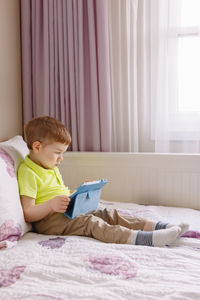 Cute boy using digital tablet while sitting on bed at home