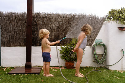 Side view of cheerful shirtless little boy pouring water from hose on sister in swimsuit while playing together in yard on summer day