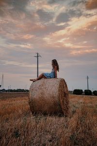 Woman standing by hay bales on field against sky during sunset