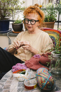 Senior woman knitting sweater while sitting at table in balcony