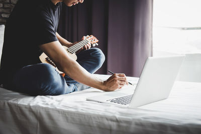 Low section of man learning guitar through laptop on bed