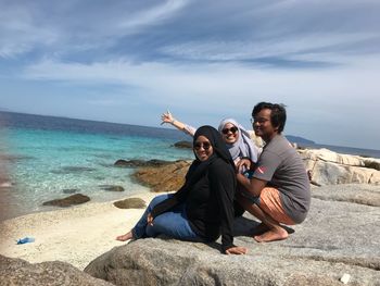 Friends sitting on rock at beach against sky