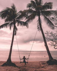 Woman on swing by palm trees at beach during sunset