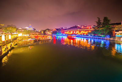 Illuminated buildings by river against sky at night