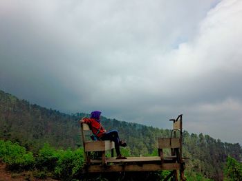 Side view of woman sitting on bench against cloudy sky