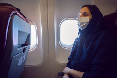 Woman in a black jacket with mask is a passenger of the plane sitting on chair next to the window