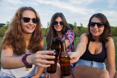 Portrait of smiling young woman toasting alcohol bottles while sitting on field