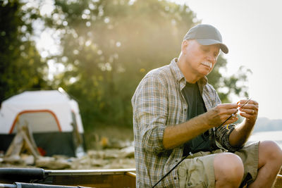 Senior man adjusting fishing tackle while sitting on boat against trees