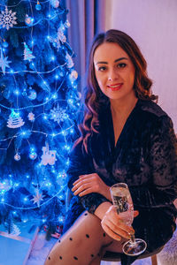 Happy young woman sitting with glass of champagne and bengal light next to the christmas tree. new 