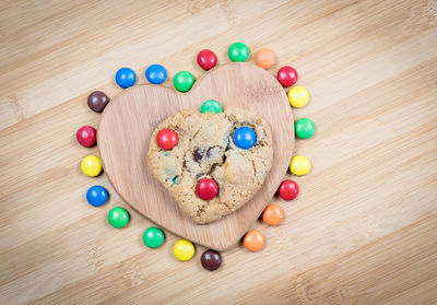 Directly above view of fresh cookie on heart shape serving board amidst colorful candies over wooden table