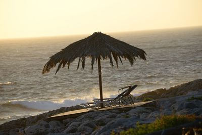 Deck chairs and parasol at sea shore against sky during sunset