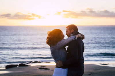 Rear view of couple standing at beach against sky during sunset