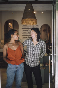 Happy multiracial female sales colleagues standing together at fashion store seen through doorway