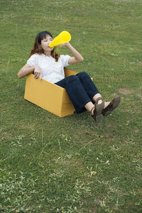 Full length of woman sitting with megaphone in box on grass