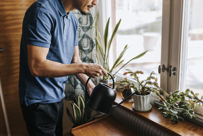 Midsection of male caregiver watering plants on window sill at home