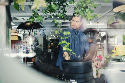 Thoughtful man looking away while working in shop seen through glass window