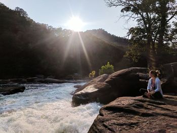 Young woman sitting on rock against sky at sunset