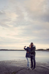 Smiling couple taking selfie while standing at beach against sky during sunset