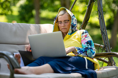 Mature woman with laptop and documents working in garden on rocking couch, green home office concept