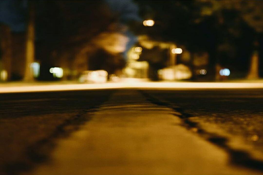 surface level, illuminated, the way forward, night, transportation, selective focus, street, diminishing perspective, road, defocused, vanishing point, empty, asphalt, road marking, focus on foreground, in a row, no people, outdoors, street light, city