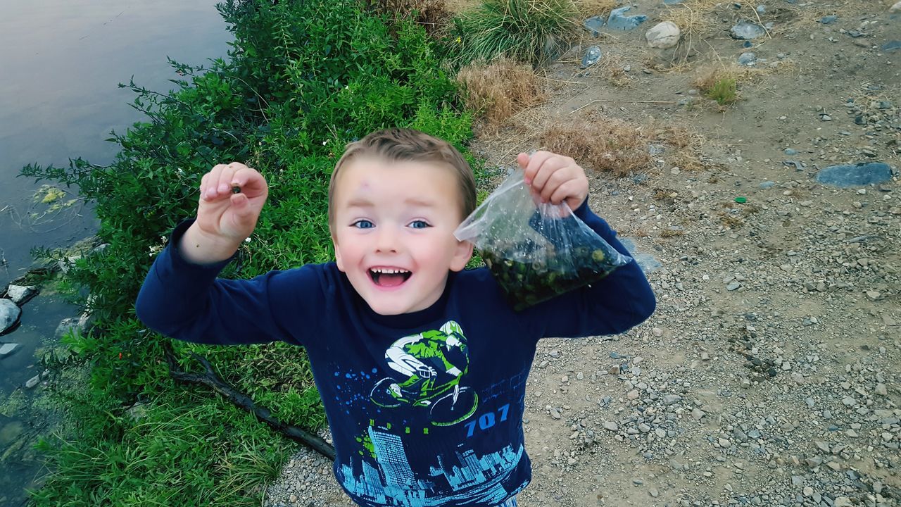 real people, looking at camera, portrait, childhood, front view, mouth open, one person, boys, elementary age, high angle view, outdoors, lifestyles, leisure activity, day, casual clothing, happiness, gesturing, smiling, standing, growth, grass, water, tree, nature