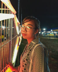 Portrait of young woman standing by fence at night