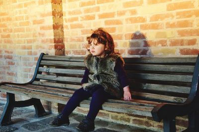 Full length of woman sitting on bench against brick wall