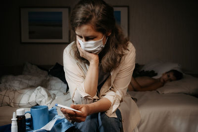 Upset and worried woman siting on bed and looking at thermometer. little child laying on bed