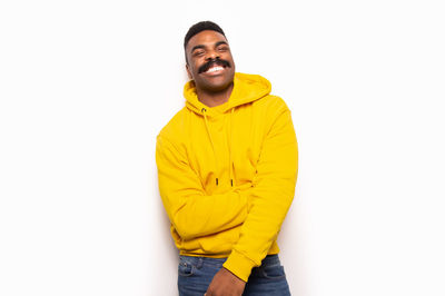 Cheerful african american man in yellow hoodie smiling and looking at camera while standing against white background