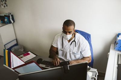 Male healthcare worker wearing face mask using computer in medical clinic