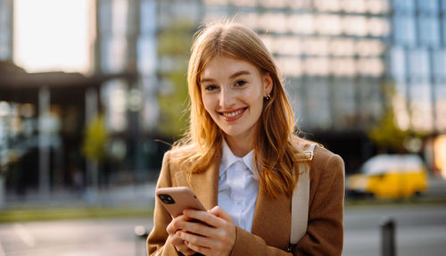 Professional in formal wear smiling for a portrait photograph with a mobile phone in a city street