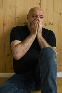 Worried mature man with hands covering mouth sitting near wooden wall at home